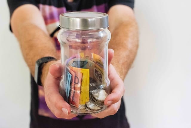 A mans arms outstretched holding a glass jar of saved Australian money