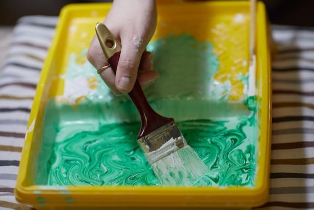 Womans hand holds paint brush and mixes white and green paint in yellow tray